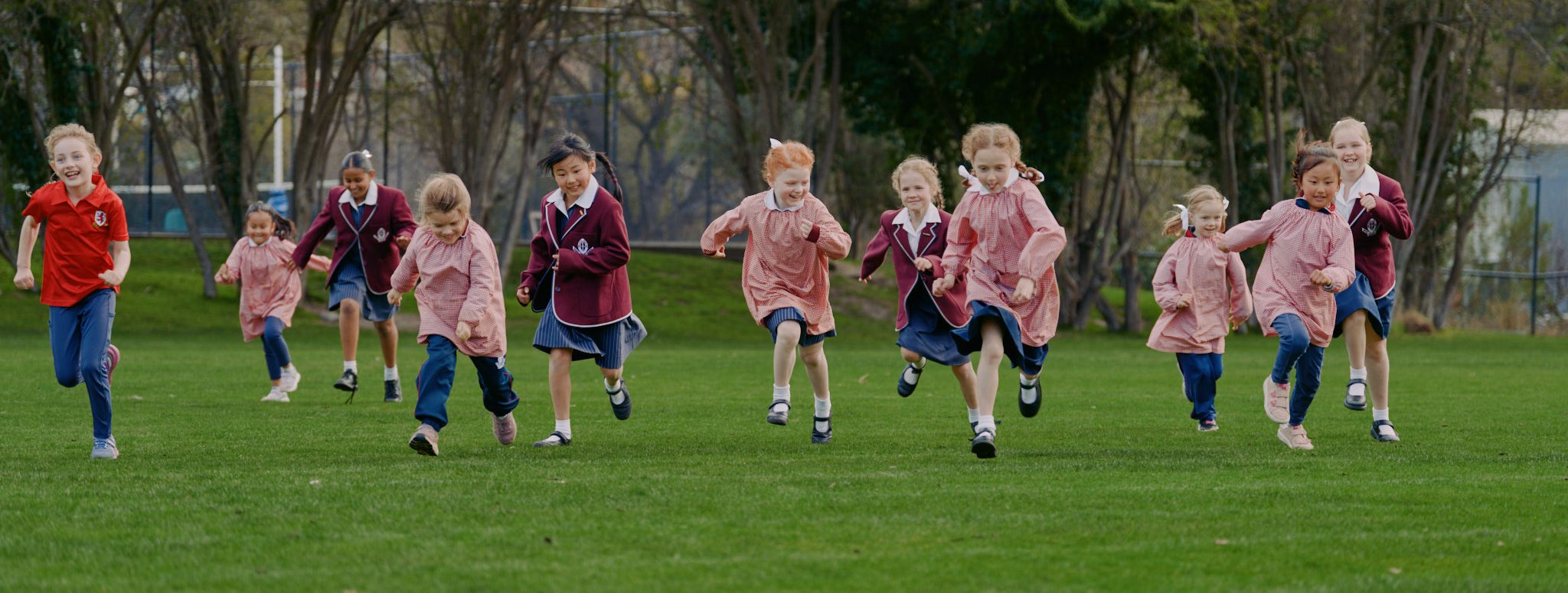 A group of laughing mixed aged Junior School students in uniform running accross the oval towards the camera.